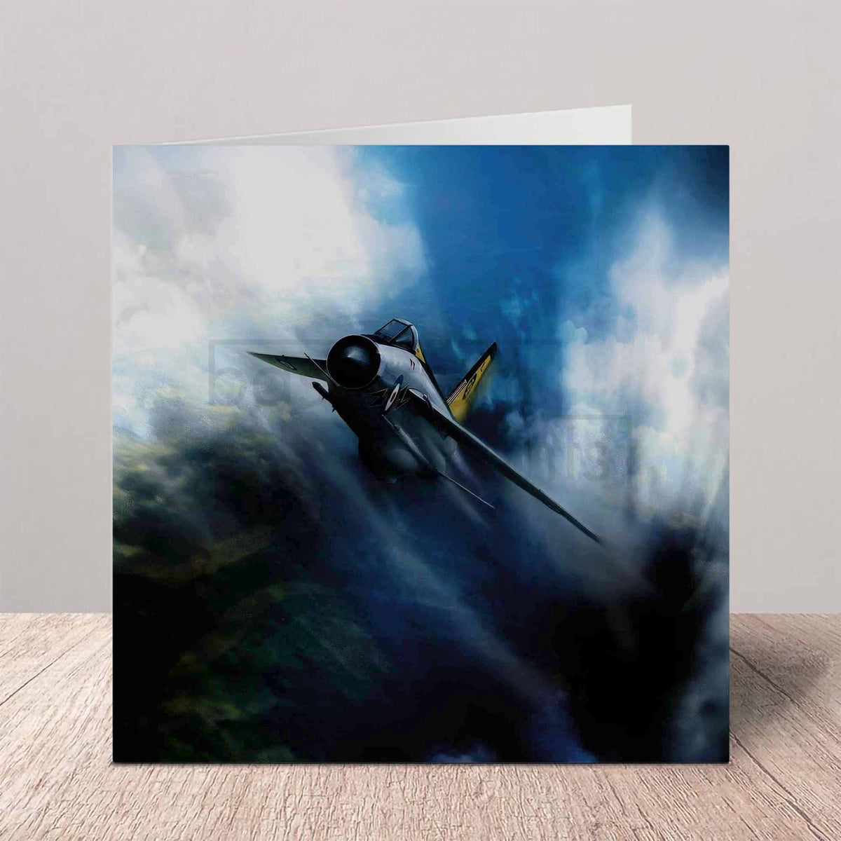 A square art print or card featuring a dramatic painting of a fighter jet flying through dark blue and white storm clouds, displayed standing on a light wood surface against a neutral gray wall.