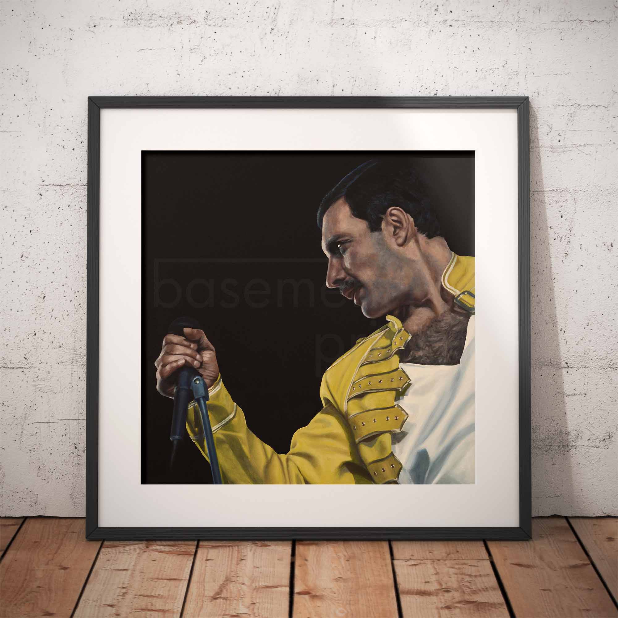 Framed portrait of Freddie Mercury in a yellow outfit against a dark background, displayed on a wooden floor with a white brick wall.