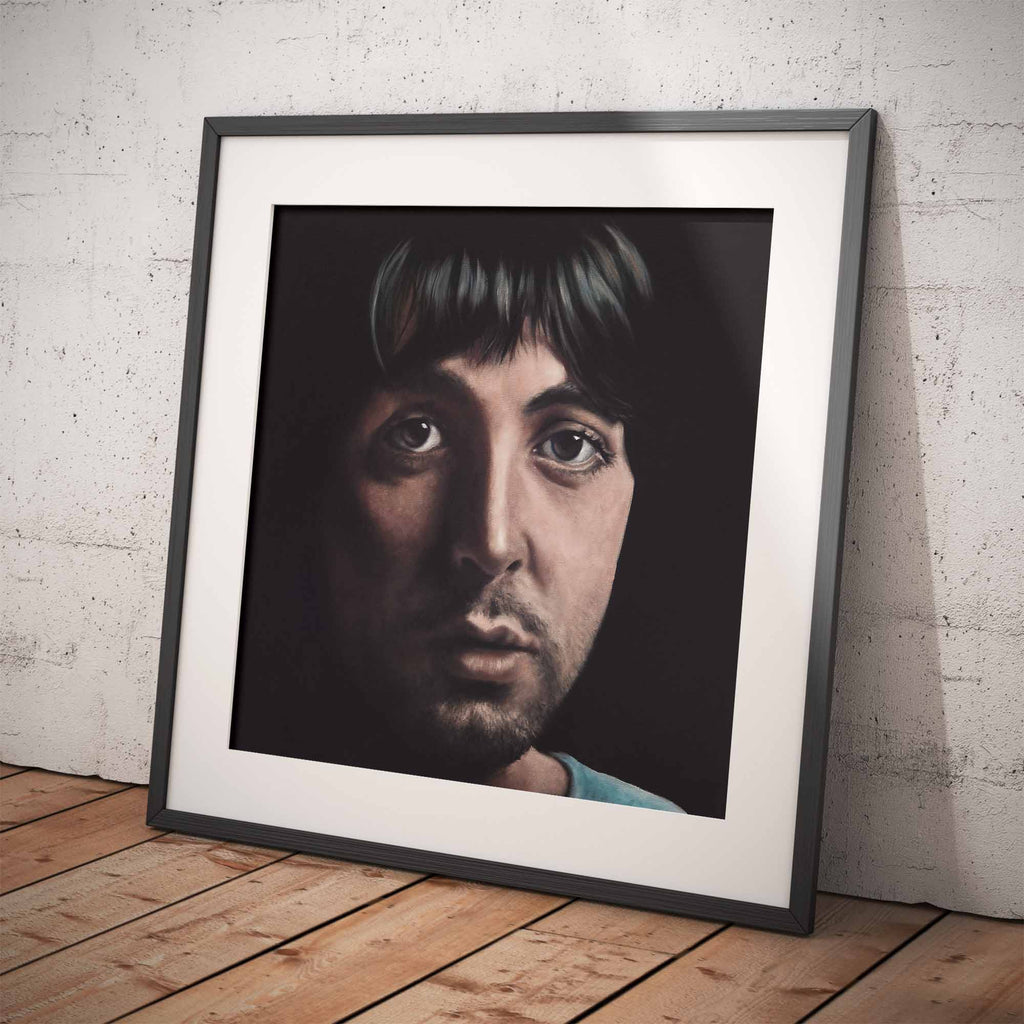 Framed portrait of Paul McCartney on a wooden floor with a white brick wall background