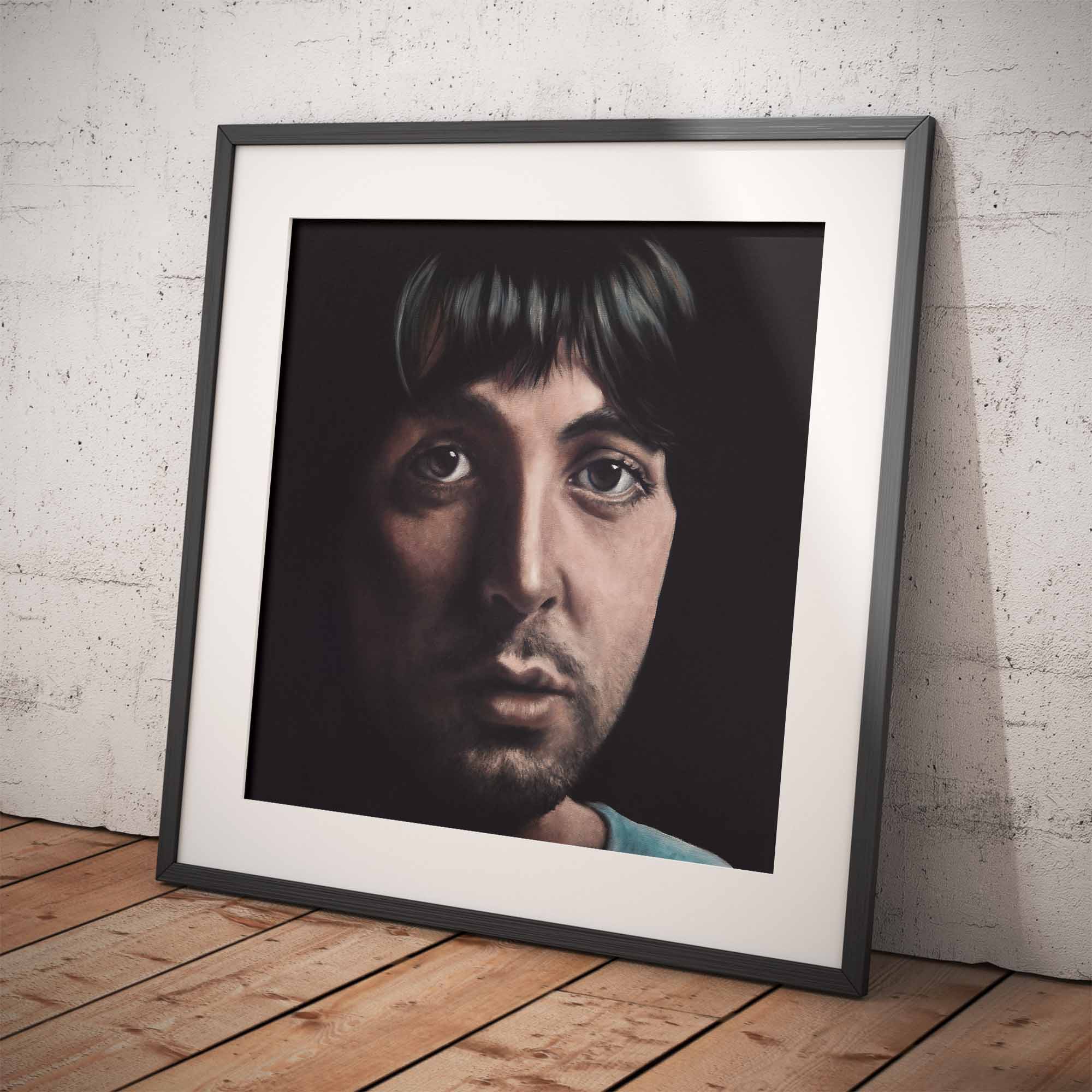 Framed portrait of Paul McCartney on a wooden floor with a white brick wall background