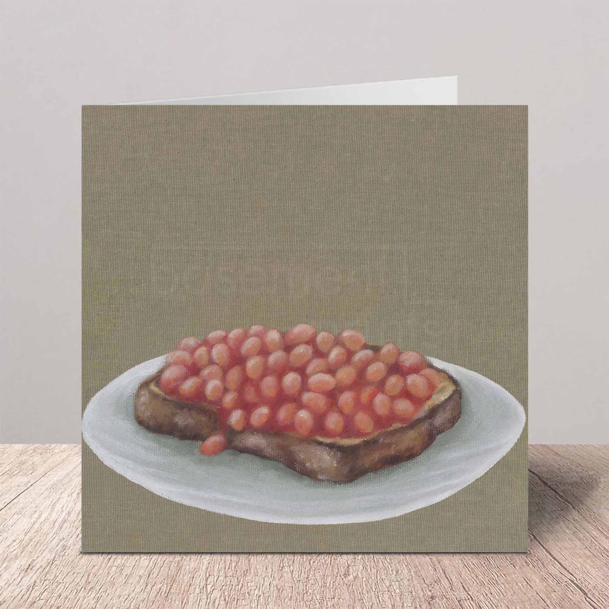A square greeting card featuring a realistic oil painting of baked beans on toast, standing on a wooden surface against a neutral background.