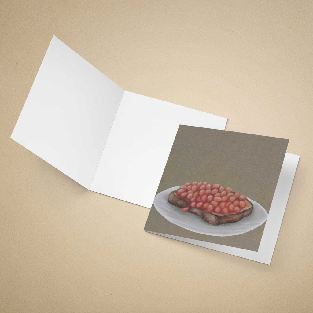 An open white greeting card lies on a beige background, with a printed card insert showing an oil painting of baked beans on toast.