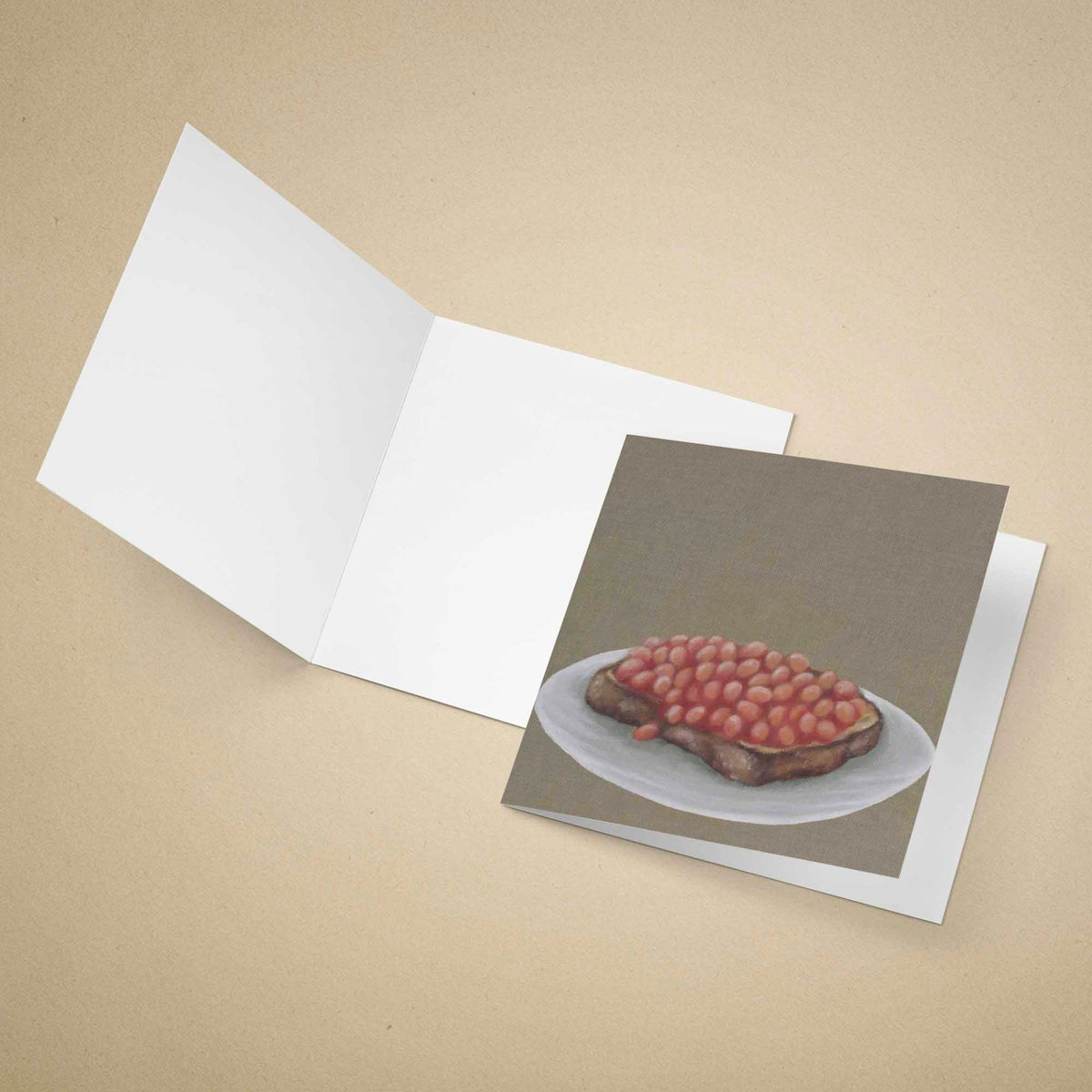 An open white greeting card lies on a beige background, with a printed card insert showing an oil painting of baked beans on toast.