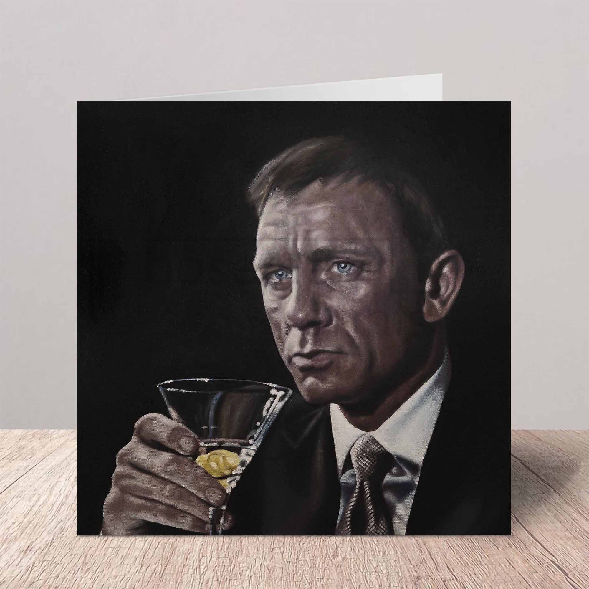 Greeting card Daniel Craig as James Bond holding a cocktail glass on a wooden surface.