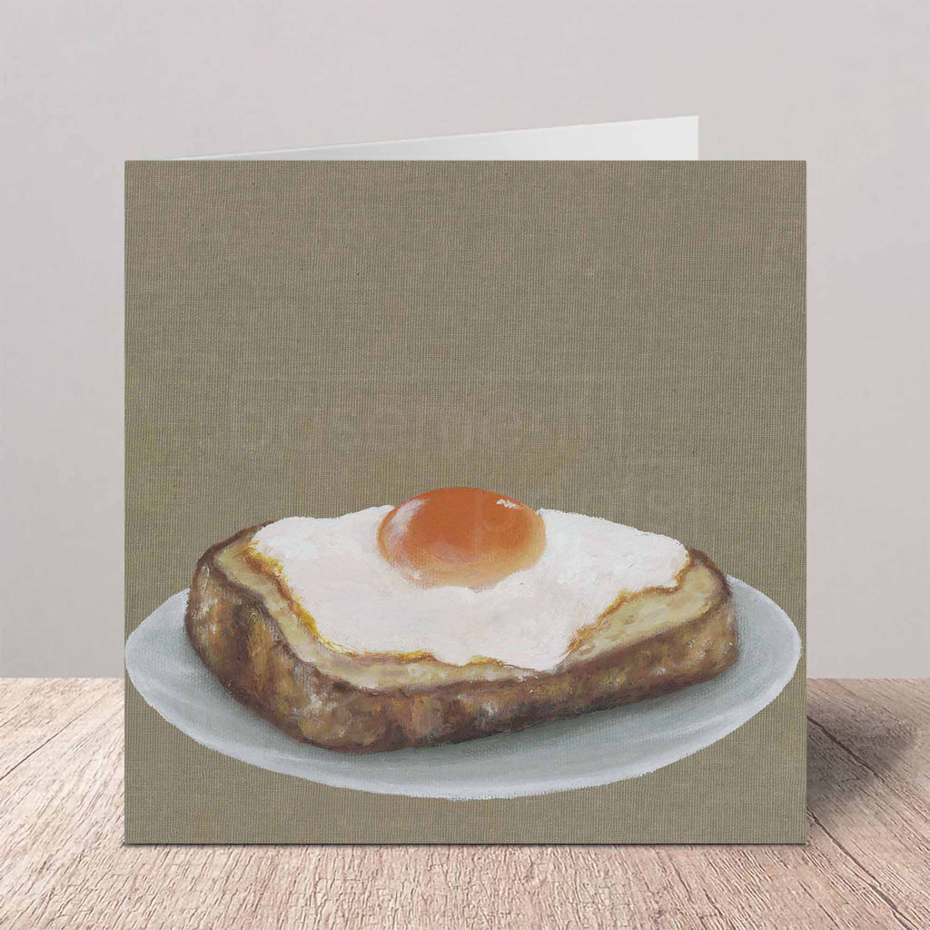 A square greeting card featuring a realistic oil painting of a fried egg on toast, standing on a wooden surface against a neutral background.