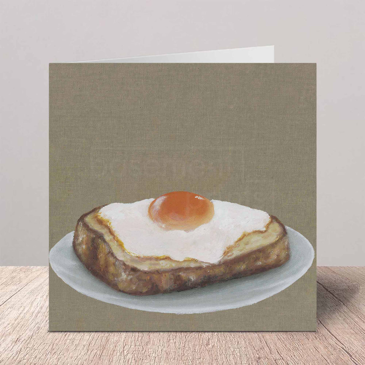 A square greeting card featuring a realistic oil painting of a fried egg on toast, standing on a wooden surface against a neutral background.