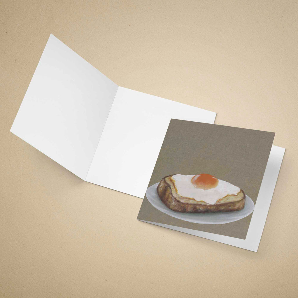 An open white greeting card lies on a beige background, with a printed card insert showing an oil painting of a fried egg on toast.