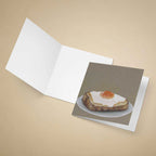 An open white greeting card lies on a beige background, with a printed card insert showing an oil painting of a fried egg on toast.