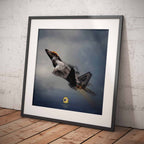Framed artwork of an F22 Raptor fighter jet against a cloudy sky on a wooden floor with white brick wall background.