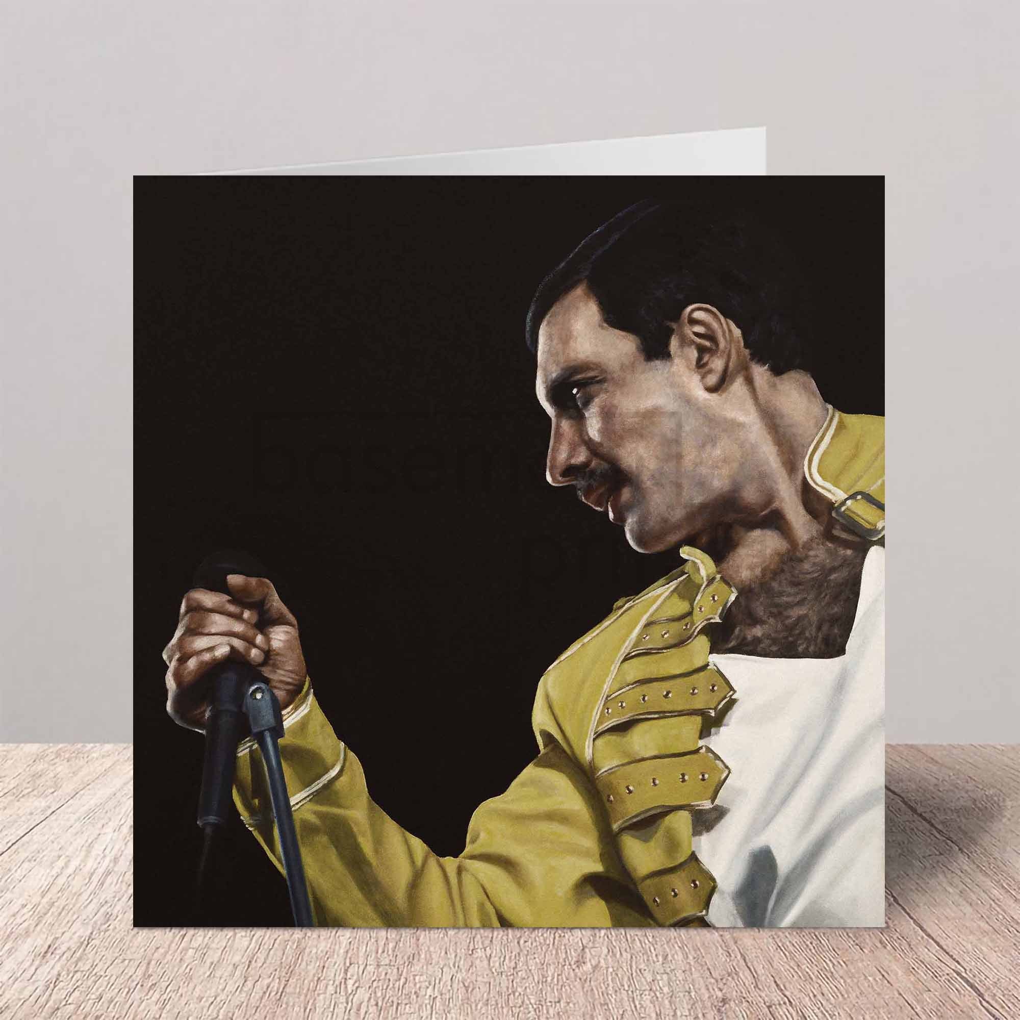 Greeting card with a portrait of Freddie Mercury in a yellow jacket holding a microphone on a wooden surface.