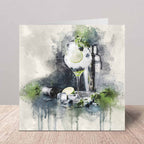 A square greeting card featuring a watercolor illustration of a gin and tonic with a lime slice, ice, mint leaves, and a shaker, standing on a wooden surface against a neutral gray background.