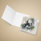An open white greeting card lies on a beige background, with a printed watercolor-style illustration on the front showing a gin and tonic glass, green leaves, and dark gray watercolor washes.