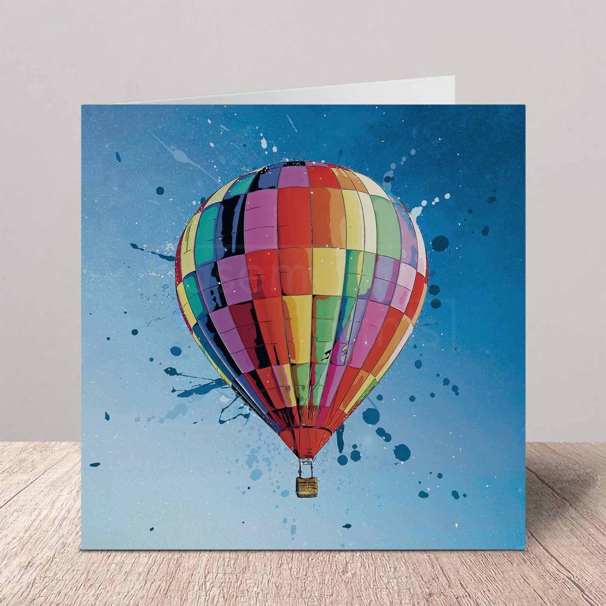 A square greeting card standing upright on a light wood surface against a neutral gray wall. The card features a colorful hot air balloon illustration with a blue sky and paint-splatter background design.