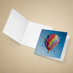 An open white greeting card on a beige background, showing a printed illustration of a colorful hot air balloon against a blue sky on the front.
