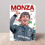 A printed card or poster featuring a stylized portrait of a Formula 1 driver celebrating at Monza, with large red text reading 'MONZA WINNER 2026' and paint-splatter graphics. The card is photographed standing upright on a light wood surface against a gray wall.