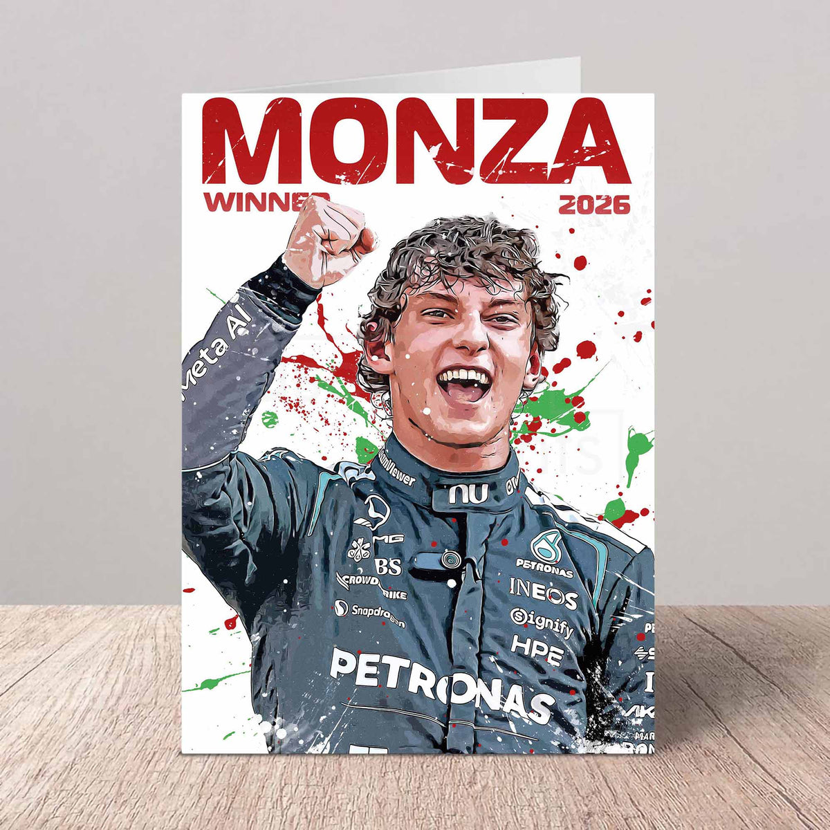 A printed card or poster featuring a stylized portrait of a Formula 1 driver celebrating at Monza, with large red text reading 'MONZA WINNER 2026' and paint-splatter graphics. The card is photographed standing upright on a light wood surface against a gray wall.