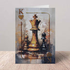 A vertical greeting card featuring an artistic painting of chess pieces, with a prominent gold king and black pawns on a textured gray and gold background. The card is displayed standing upright on a light wood surface against a neutral gray wall.