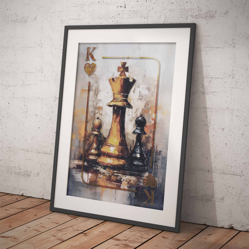 A framed art print leaning against a textured light gray wall on a wooden floor. The print features a stylized painting of chess pieces, with a gold king and black pieces on a muted gray and gold background.