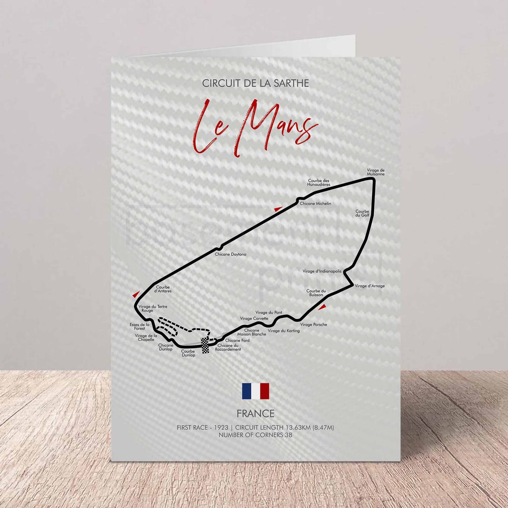 A printed greeting card featuring a black race track map of Le Mans with the text 'Circuit de la Sarte Le Mans' and 'France' at the bottom, standing upright on a light wood surface against a neutral wall.
