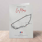 A printed greeting card featuring a black race track map of Le Mans with the text 'Circuit de la Sarte Le Mans' and 'France' at the bottom, standing upright on a light wood surface against a neutral wall.