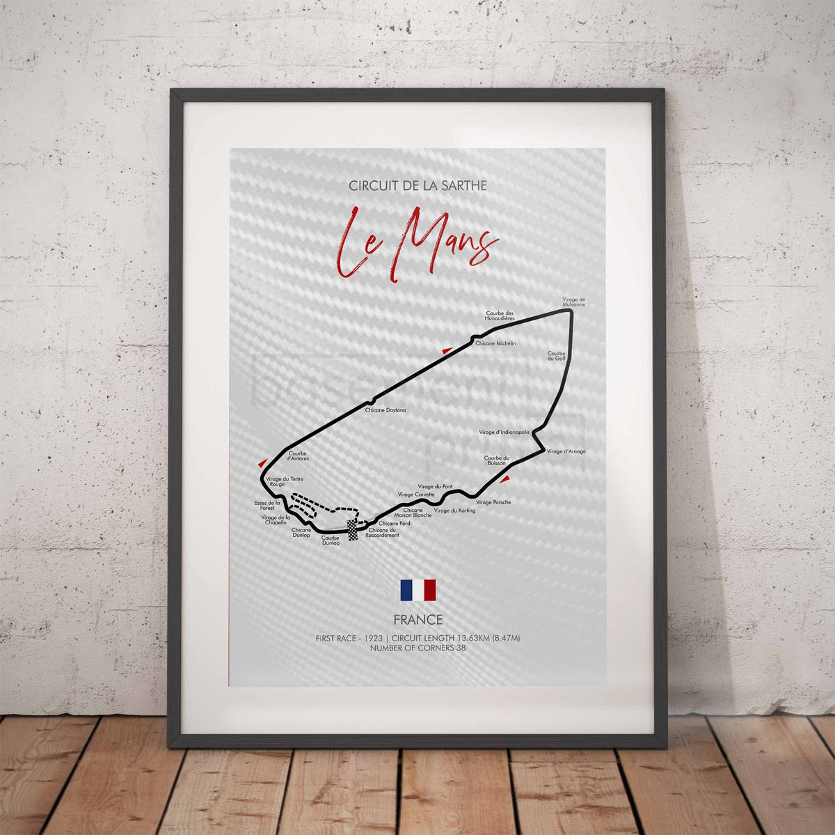 A framed poster print of the Le Mans race track in France, featuring a black line map on a light gray patterned background with red script text. The framed print is displayed leaning against a white textured wall on a wooden floor.