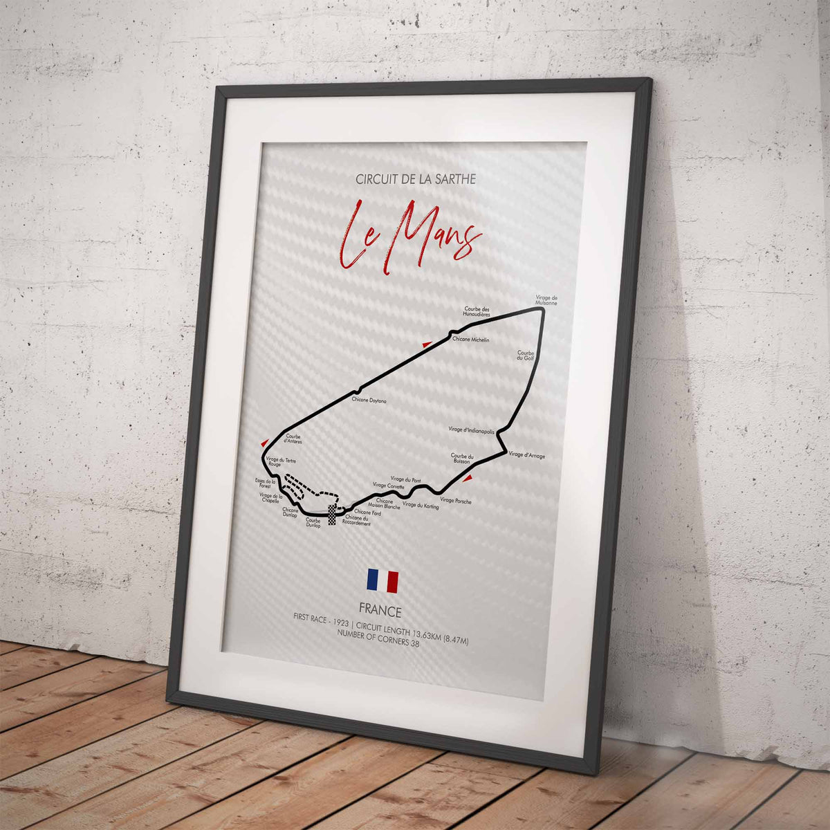 A framed print of the Le Mans race track circuit map of France, leaning against a textured white wall on a wooden floor.