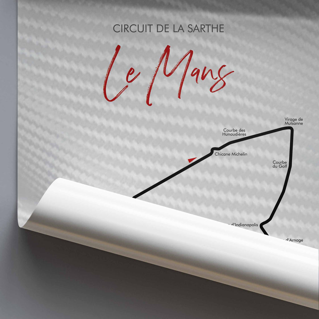 A rolled-up poster or print showing the Circuit de la Sarthe Le Mans race track, with a large red script title 'Le Mans' and black track-line illustration on a light gray background.