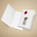 Greeting card with a light brown Cockapoo holding a red balloon on a beige background