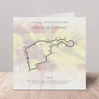 A square printed card or poster featuring a Formula 1 Spanish Grand Prix circuit map of Circuit de Madring, with track numbers, course details, and a soft pastel watercolor background, displayed on a wooden surface against a light gray wall.