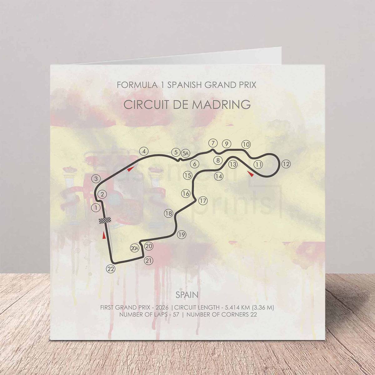 A square printed card or poster featuring a Formula 1 Spanish Grand Prix circuit map of Circuit de Madring, with track numbers, course details, and a soft pastel watercolor background, displayed on a wooden surface against a light gray wall.