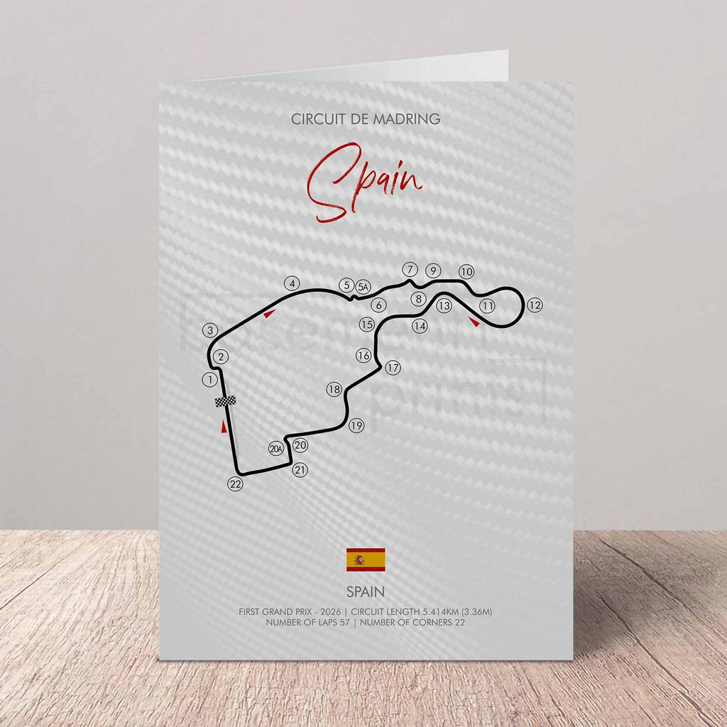 A printed circuit map card for Circuit de Madrid, Spain, standing on a light wood surface against a neutral gray background. The card features a white and gray carbon-fiber textured pattern, the circuit track outline in black with red Arrow markers, and a small Spanish flag near the bottom.