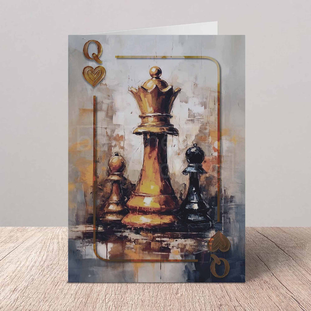 A vertical greeting card featuring an artistic painting of chess pieces, with a prominent gold queen and black pawns on a textured gray and gold background. The card is displayed standing upright on a light wood surface against a neutral gray wall.