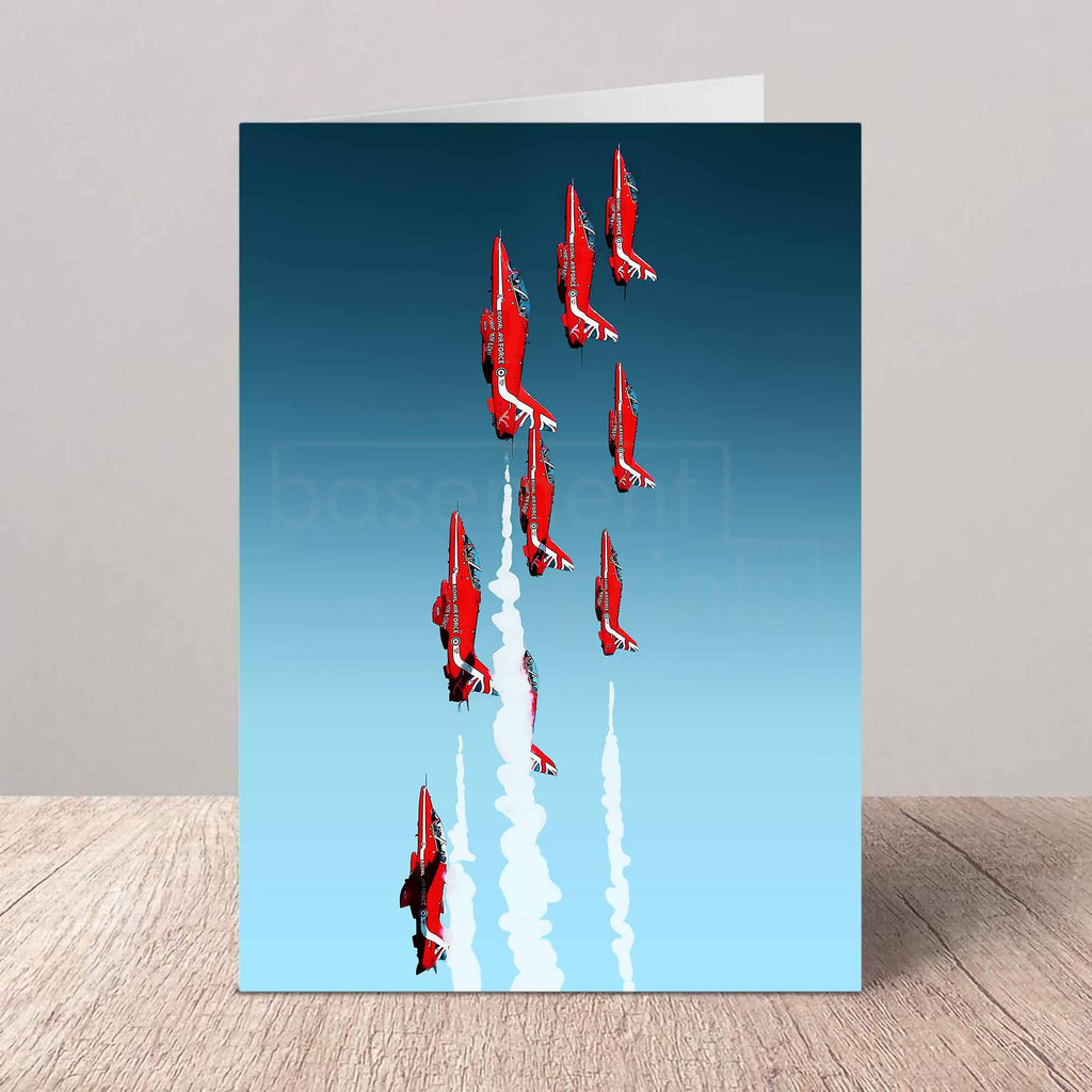 A vertical greeting card standing on a light wood surface against a neutral gray wall. The card features a graphic illustration of nine Red Arrows jets flying in formation and leaving white smoke trails against a blue-to-light-blue gradient sky.