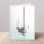 A folded greeting card standing upright on a light wood surface against a neutral gray wall. The card features a printed photograph of three sailboats on calm water under a pale sky.