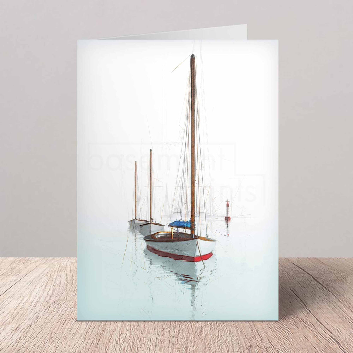 A folded greeting card standing upright on a light wood surface against a neutral gray wall. The card features a printed photograph of three sailboats on calm water under a pale sky.