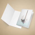An open white greeting card on a warm beige background, with the front cover showing a printed photograph of a sailboat on calm water.