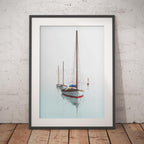 A framed art print depicting three sailboats on a calm, misty light blue water surface, leaning against a white textured brick wall on a wooden floor.