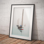 A framed art print of a classic sailboat on calm water, leaning against a white brick wall on a wooden floor.