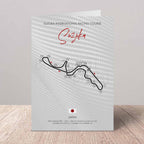 A vertical printed greeting card featuring a stylized Suzuka International Racing Course map with numbered turns, red script title 'Suzuka,' and course text, standing on a wooden surface against a light neutral wall.