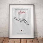 A framed racing course poster for Suzuka International Racing Course, leaning against a white textured wall on a wooden floor. The poster features a light gray patterned background with a black track line, red script title 'Suzuka'.