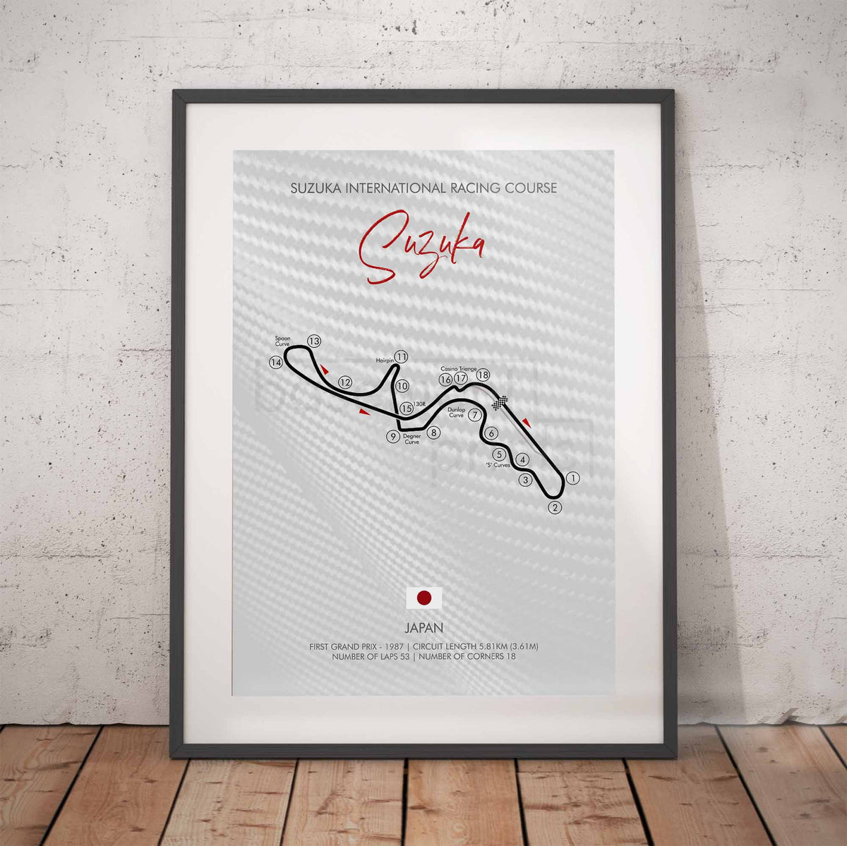 A framed racing course poster for Suzuka International Racing Course, leaning against a white textured wall on a wooden floor. The poster features a light gray patterned background with a black track line, red script title 'Suzuka'.