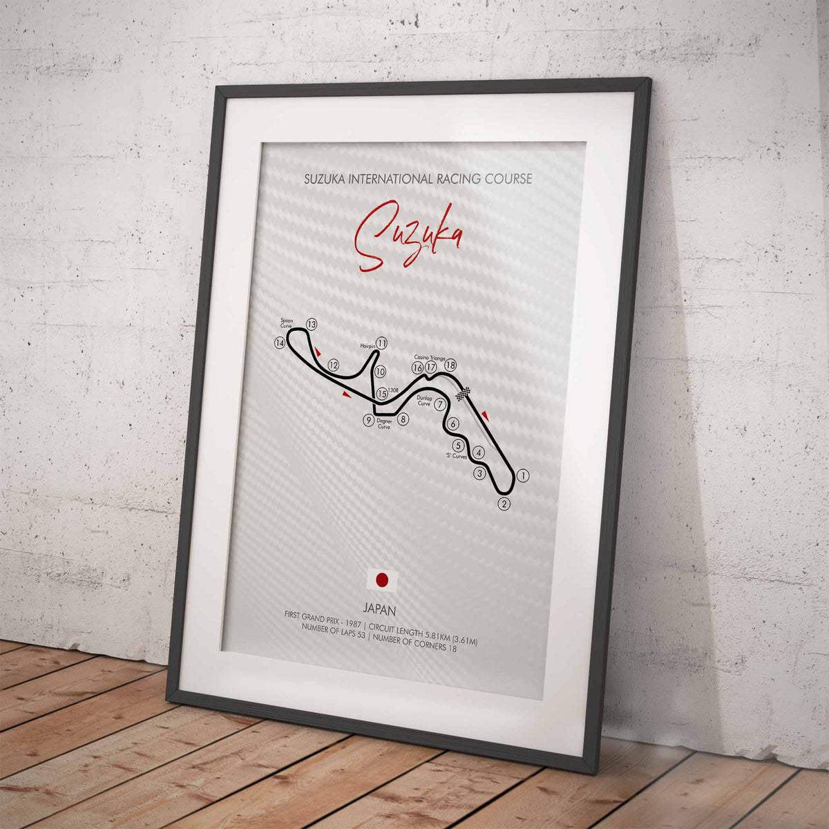 A framed Suzuka International Racing Course poster leaning against a textured light gray wall on a wooden floor. The poster has a black frame, white mat, and a light gray printed background with a black track map, red script title 'Suzuka', and small red location markers.