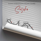 A rolled poster displaying a printed Suzuka International Racing Course map and track layout on a light gray background with a carbon-fiber texture.