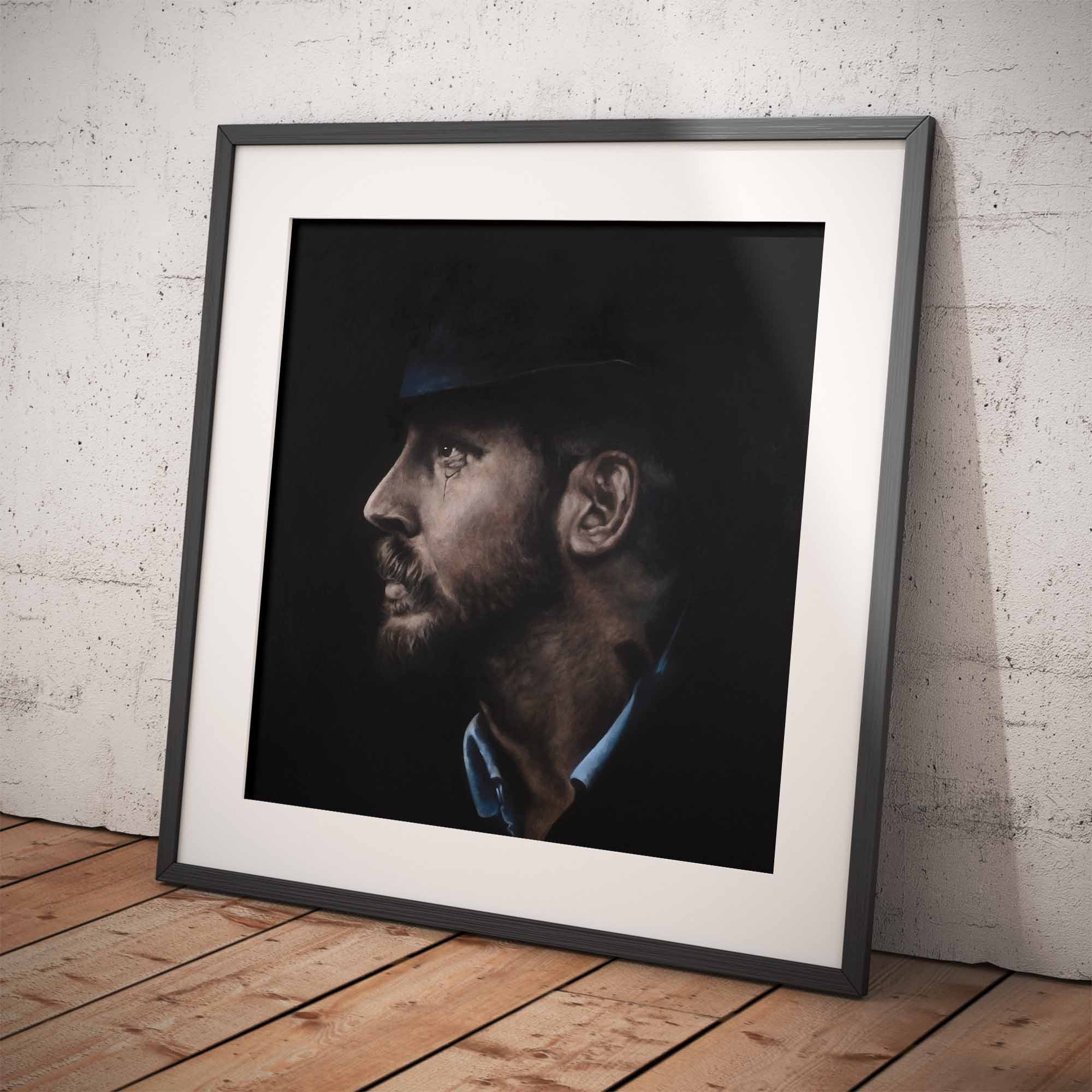 Framed portrait of a Tom Hardy on a wooden floor against a white brick wall.